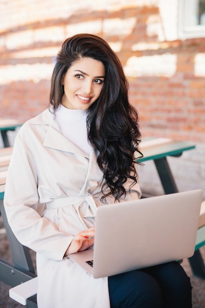 Modisches weibliches Model im eleganten weißen Mantel, das auf einer Bank sitzt und einen Laptop hält, der in sozialen Netzwerken surft Professionelle Geschäftsfrau, die im Café im Freien sitzt und mit einem modernen Computer arbeitet