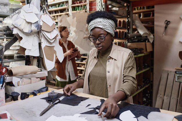 Modista africana cortando los trozos de tela para coser ropa en la mesa mientras trabajaba en la fábrica