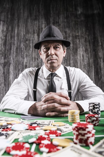 Möchten Sie spielen? Ernster älterer Mann in Hemd und Hosenträgern, der am Pokertisch sitzt und mit Karten mit Geld und Spielchips in die Kamera schaut, die überall um ihn herum liegen