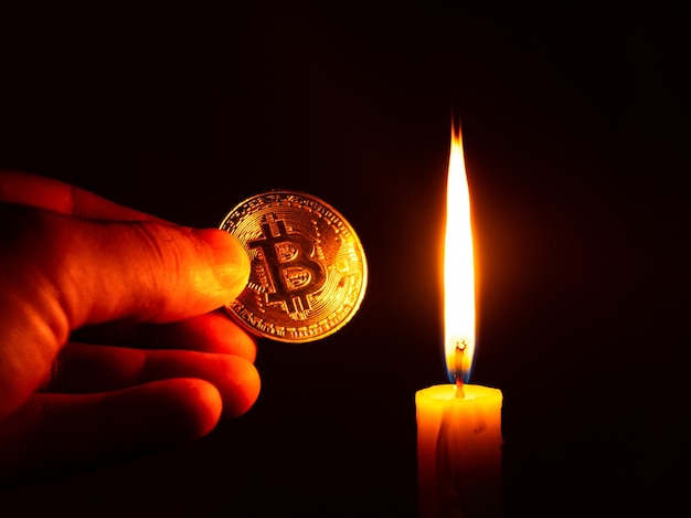 Moeda de ouro bitcoin na mão sob a luz quente de uma vela em um fundo escuro, dinheiro virtual, conceito de criptomoeda.