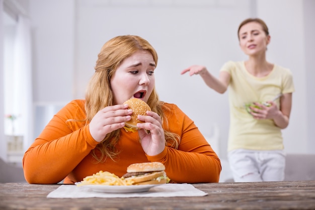 Mofa. Infeliz mujer corpulenta comiendo un sándwich y su delgada amiga criticandola