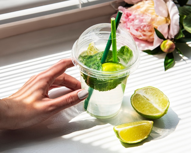 Mojito-Cocktail in weiblichen Händen vor dem Fenster und ein Strauß Pfingstrosen