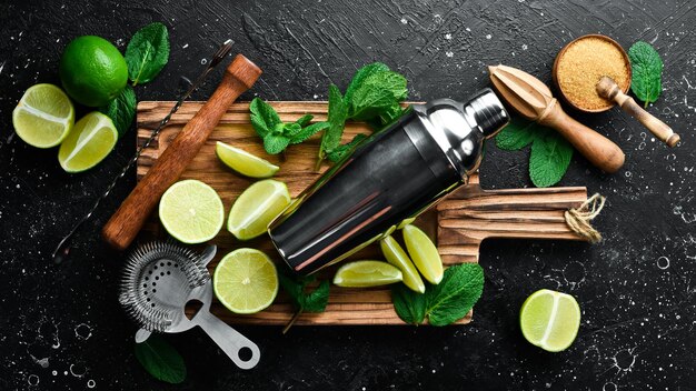 Mojito-Shaker, Zucker, frische Limette und Minze auf Holzbrett auf schwarzem Steinhintergrund, Draufsicht