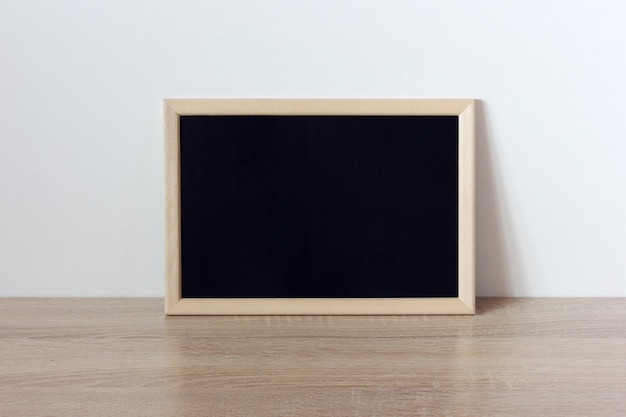 Moldura de madeira com quadro negro de fundo em branco em uma prateleira de mesa contra uma maquete de parede branca de volta às aulas