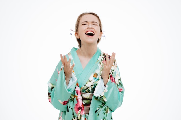 Molesta y frustrada joven geisha en kimono japonés tradicional gritando y gritando levantando los brazos de pie sobre la pared blanca