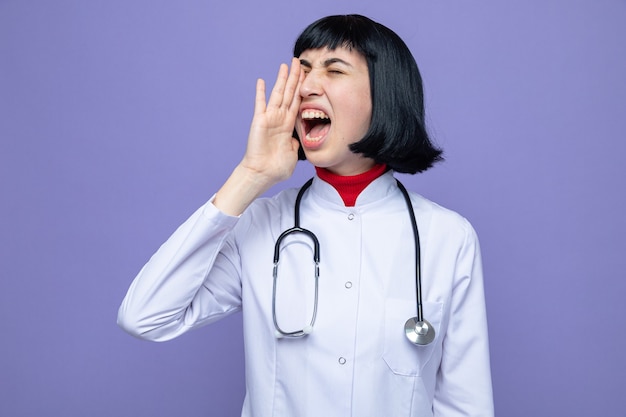 Molesta joven bastante caucásica en uniforme médico con estetoscopio sosteniendo la mano cerca de la boca llamando a alguien