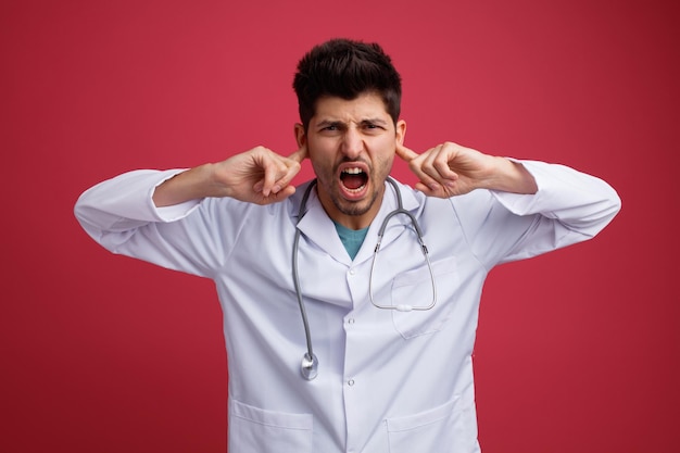 Molesto joven médico masculino vistiendo uniforme médico y estetoscopio poniendo los dedos en los oídos mirando a la cámara aislada sobre fondo rojo.