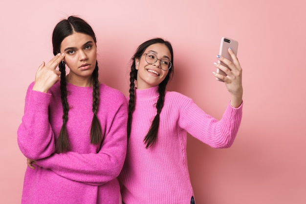 Molesto ofendido adolescente de pie junto a su feliz amiga con teléfono celular aislado sobre pared rosa