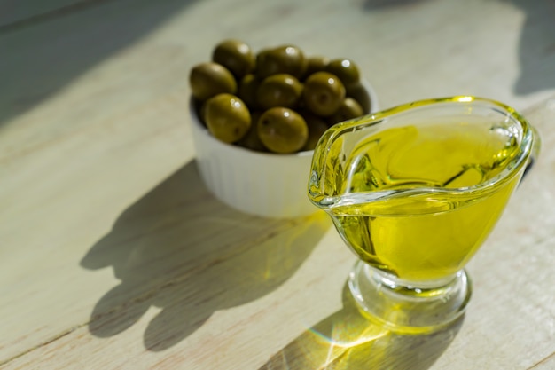 Molheira de vidro com azeite de oliva extra virgem e azeitonas verdes frescas na mesa de madeira.