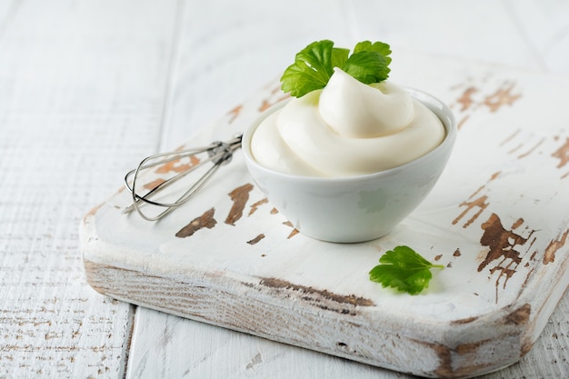 Molho de maionese tradicional em tigela de cerâmica branca e ingredientes para sua preparação em madeira branca.