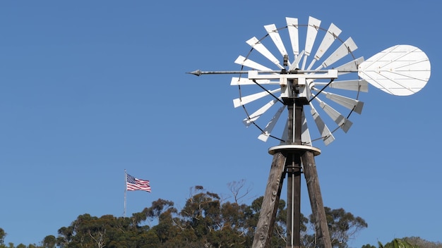 Molino de viento, rotor de palas y bandera de Estados Unidos contra el cielo azul. Turbina eólica con bomba de agua, generador de energía.