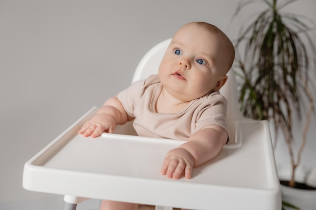 Molliges Baby in einem weißen Body sitzt in einem weißen Hochstuhl zum Füttern