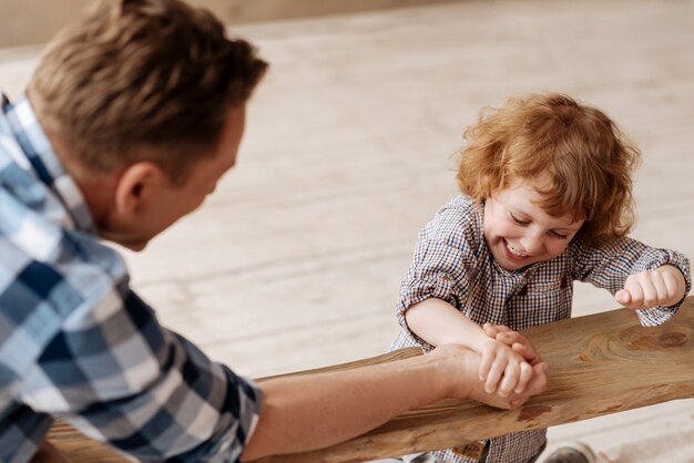 Moment des Glücks. Positiv entzückter Junge, der ein Lächeln auf seinem Gesicht behält und seine Hand beugt, während er mit Vater spielt