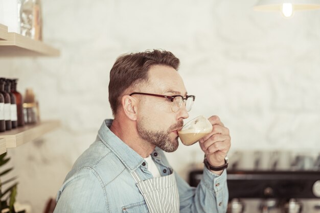 Momento de apreciação. Homem bonito na cozinha bebendo seu cappuccino e sorrindo.