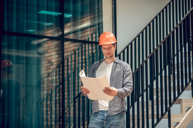 Momento de trabalho. Homem adulto jovem atento envolvido no capacete de segurança, olhando para o desenho no papel nas mãos, em pé no novo edifício moderno