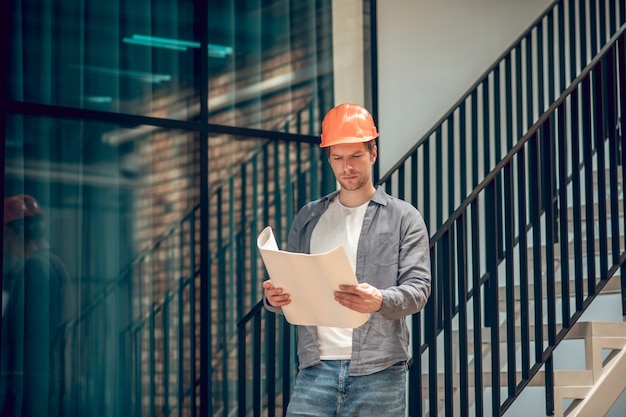 Momento de trabalho. homem adulto jovem atento envolvido no capacete de segurança, olhando para o desenho no papel nas mãos, em pé no novo edifício moderno