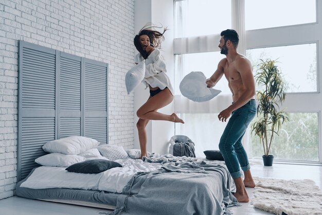 Momento divertido. Longitud total de la feliz pareja joven que tiene una divertida pelea de almohadas mientras pasa tiempo sin preocupaciones en el dormitorio