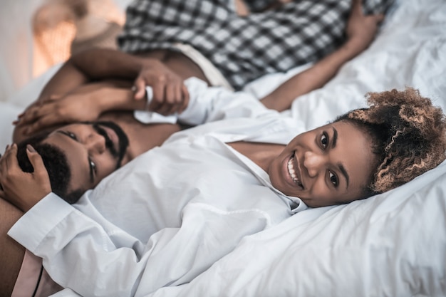 Momento feliz. Alegre joven mujer afroamericana brillante en ropa de casa abrazando a marido en la cama de buen humor