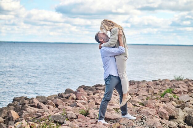 Momento feliz. Entusiasta hombre barbudo adulto levantando en sus brazos alegre mujer rubia en la orilla del mar en buen día