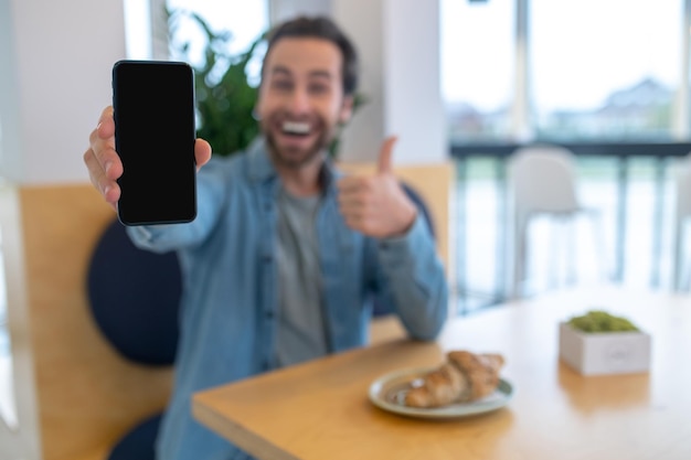 Momento feliz. Hombre mostrando la pantalla del teléfono inteligente en la cámara y gesticulando bien sentado en la mesa en el café durante el día