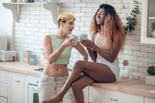 Momento feliz. Riendo linda esbelta mujer de piel oscura y novia caucásica con pelo corto teñido sosteniendo tazas en la cocina