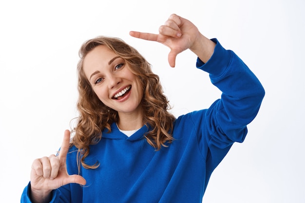 Momento positivo de captura de garota, fazendo gestos de câmera com quadros de mão e sorrindo feliz, olhando através, procurando o ângulo perfeito, em pé sobre uma parede branca