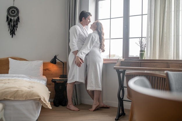 Momento romántico. Un hombre en una bata de baño blanca abrazando a una mujer cerca de la ventana.