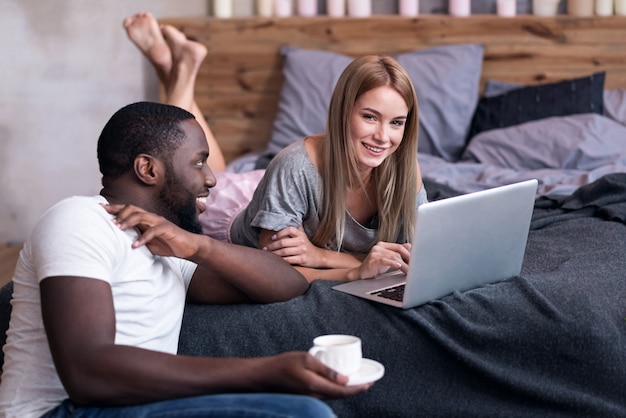 Momentos doces. Jovem casal internacional encantado, passando algum tempo juntos no quarto enquanto usa o laptop e sorrindo.