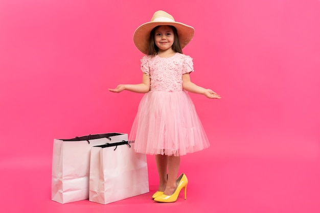 Momentos encantadores felices del tiempo de compras con la niña linda en el vestido que se coloca en los zapatos grandes de la madre con los paquetes blancos en las manos aisladas en el fondo rosa.