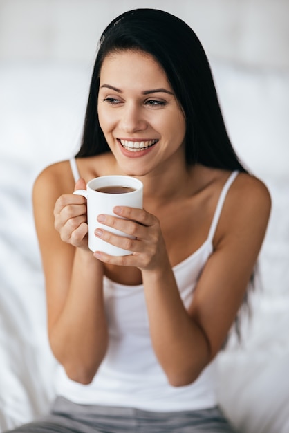 Momentos de felicidad. Hermosa mujer joven con cabello largo oscuro sosteniendo una taza y sonriendo