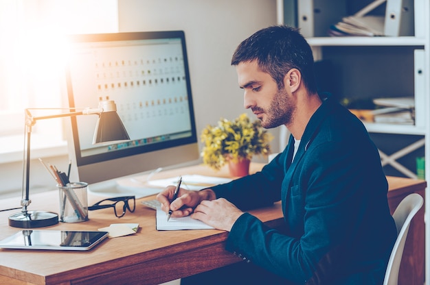 Momentos de trabajo. Vista lateral del joven confiado que escribe algo en su cuaderno mientras está sentado en su lugar de trabajo en la oficina