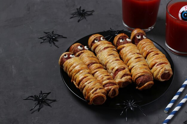 Momias de salchicha espeluznantes y jugo de tomate para la fiesta de Halloween en un plato oscuro. Idea divertida de comida para niños. Espacio para texto.