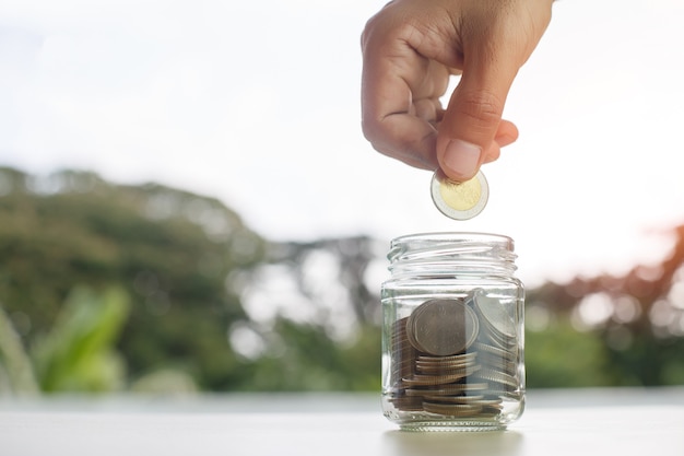 Monedas en botella de vidrio puestas sobre la mesa con la mano poniendo dinero monedas naturaleza fondo, ahorro de crecimiento o concepto de inversión.