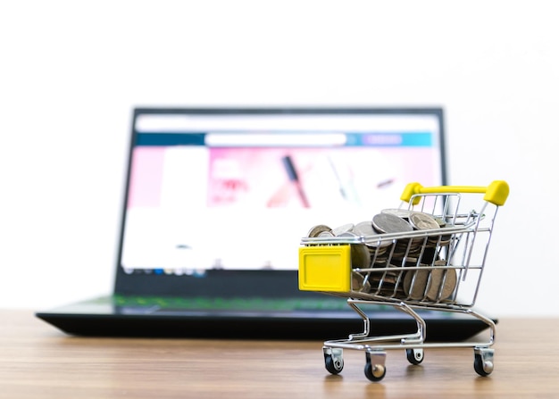 Monedas en el carrito de compras con computadora portátil en la mesa contra un fondo blanco