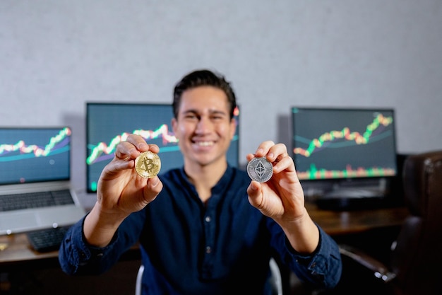 Monedas de representación física de primer plano ethereum y bitcoin. Hombre de negocios sonriente joven en el fondo con la computadora que muestra tablas y gráficos. Inversión segura en concepto de moneda criptográfica.