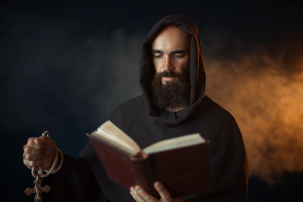 Monge medieval orando com o livro nas mãos na igreja, ritual secreto. Frade misterioso com capa escura. Mistério e espiritualidade