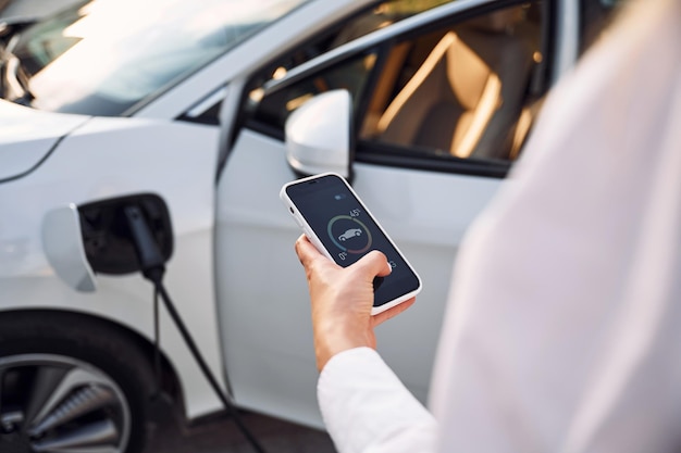 Monitoramento de carregamento por smartphone Jovem mulher com roupas brancas está com seu carro elétrico durante o dia