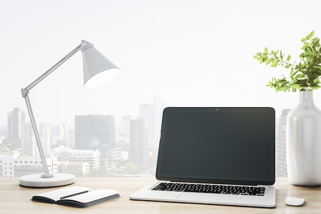 Monitore de portátil moderno en blanco en una mesa de madera con un cuaderno de lámparas elegante y un jarrón de vidrio blanco en el fondo de la vista de la ciudad Mockup