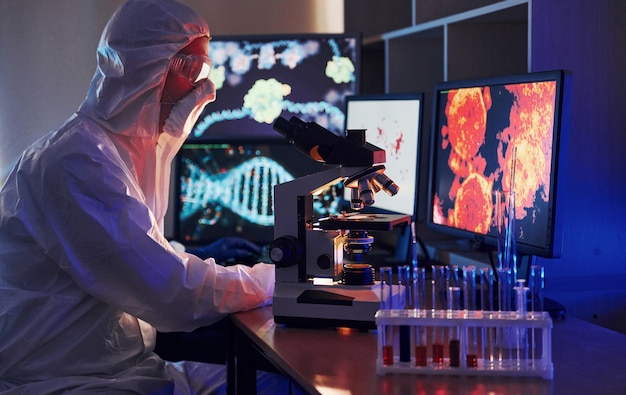 Monitores con información sobre la mesa. Científico en uniforme protector blanco trabaja con coronavirus y tubos de sangre en laboratorio