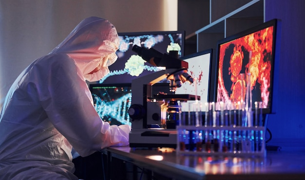 Monitores con información sobre la mesa. Científico en uniforme protector blanco trabaja con coronavirus y tubos de sangre en laboratorio