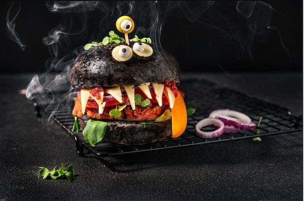 Foto monsterburger mit schwarzem brötchen, veganem patty, käse, olivenaugen und rauch auf schwarzem hintergrund. halloween-essen