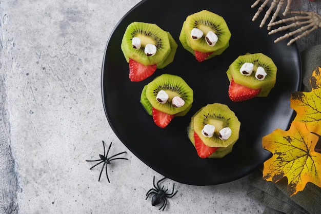 Monstros de kiwi verde assustador para Halloween Frutas Saudáveis Halloween Deleites Festa de Halloween kiwi morango maçã e monstro marshmallow em pedra cinza ou fundo de mesa de concreto Foco seletivo