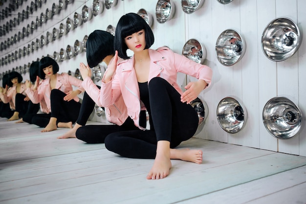 Monstruo de la moda. Chica sintética glamour, muñeca falsa con aspecto vacío y cabello negro corto está sentada en el estudio. Elegante mujer hermosa en chaqueta rosa cerca de bombillas. Concepto de moda y belleza