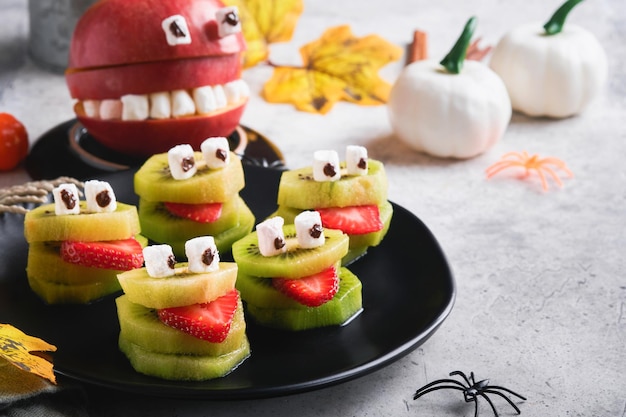 Monstruos de kiwi verde espeluznante para Halloween Fruta saludable Halloween Golosinas Fiesta de Halloween kiwi fresa manzana y monstruo de malvavisco en piedra gris o fondo de mesa de hormigón Enfoque selectivo