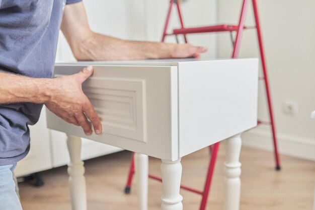 Montaje de mesita de noche blanca de madera en casa, primer plano de la mano de un trabajador haciendo muebles con herramientas profesionales. Renovación, muebles nuevos, proceso de carpintería de trabajo