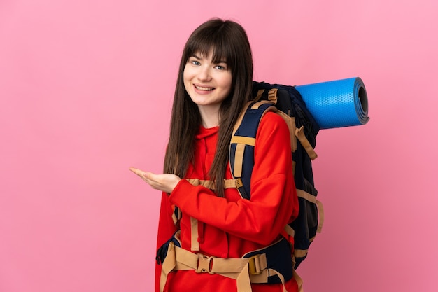 Montañero niña ucraniana con una gran mochila aislada en la pared rosa extendiendo las manos hacia el lado para invitar a venir