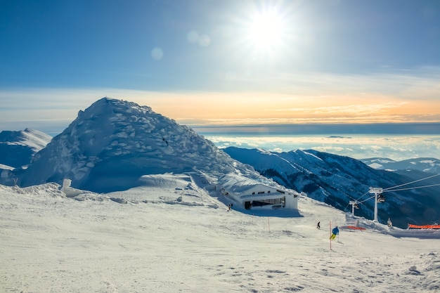 Montanhas de inverno. Picos cobertos de neve e neblina nos vales. Sol brilhante no céu azul ao longo da pista de esqui. Teleférico e bar