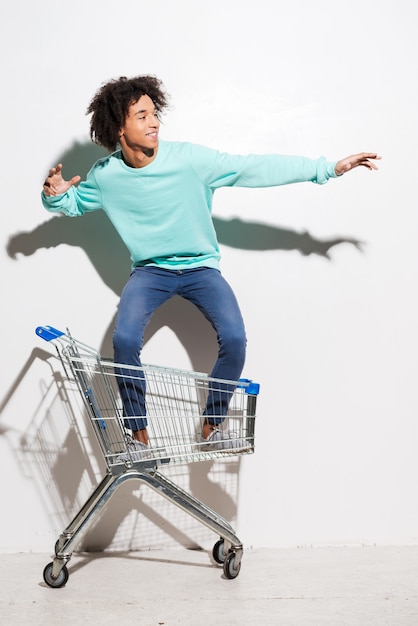 Montar en un carrito de compras. Juguetón joven africano cabalgando en carro de compras contra el fondo gris