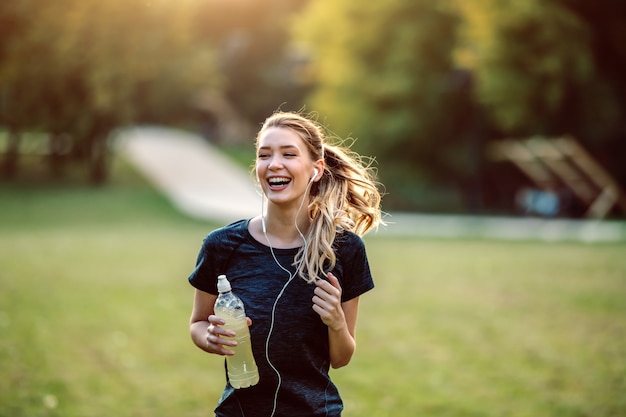 Montar deportista caucásica saludable en ropa deportiva, con cola de caballo y auriculares con refresco en las manos corriendo en la naturaleza.