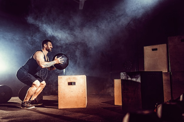 Montar un hombre barbudo tatuado saltando sobre una caja como parte de la rutina de ejercicios. Hombre haciendo salto de caja en el gimnasio. El atleta está realizando saltos de caja